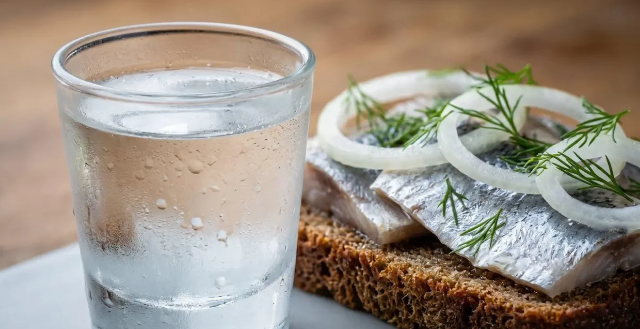 Close-up of traditional Danish akvavit shot glass beside pickled herring open-faced sandwich with onion rings