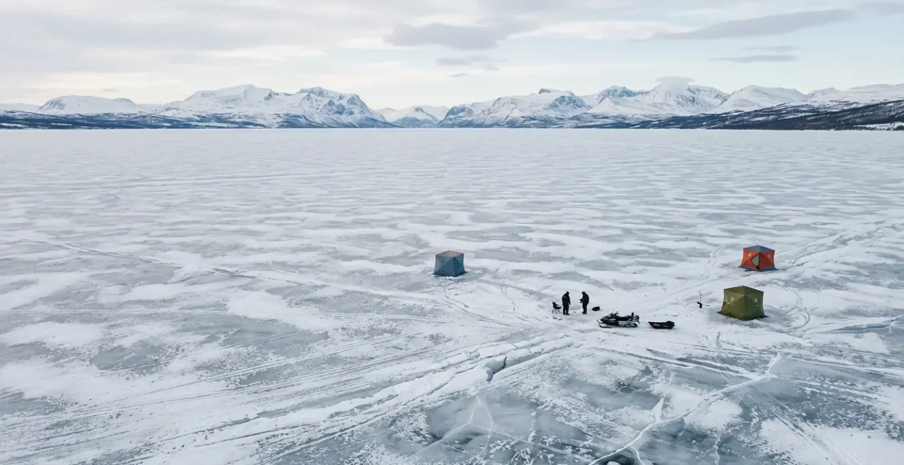 Wide aerial view of a frozen Nordic lake with scattered ice fishing shelters and distant mountains under soft winter light