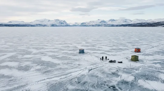 Wide aerial view of a frozen Nordic lake with scattered ice fishing shelters and distant mountains under soft winter light